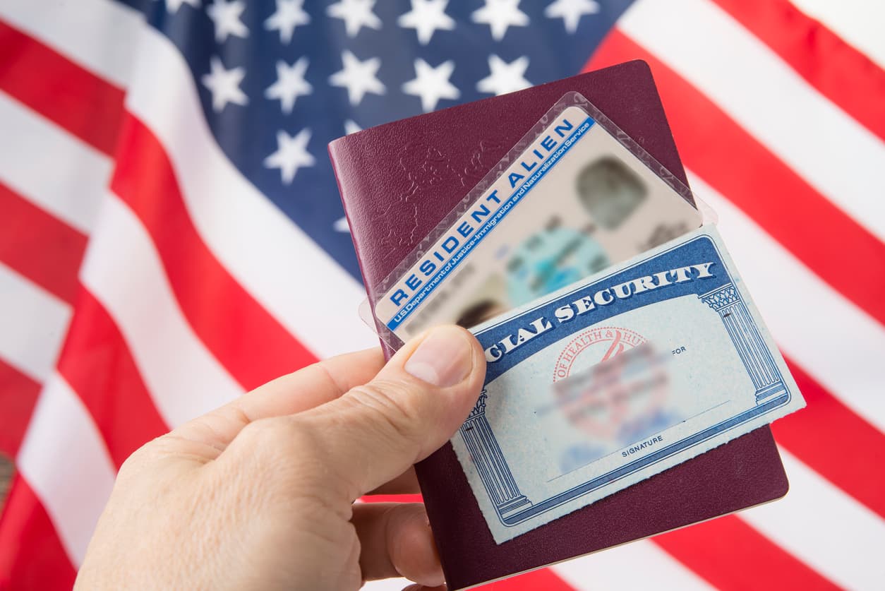 The image shows a person holding a U.S. Resident Alien card and a Social Security card, with an American flag in the background.