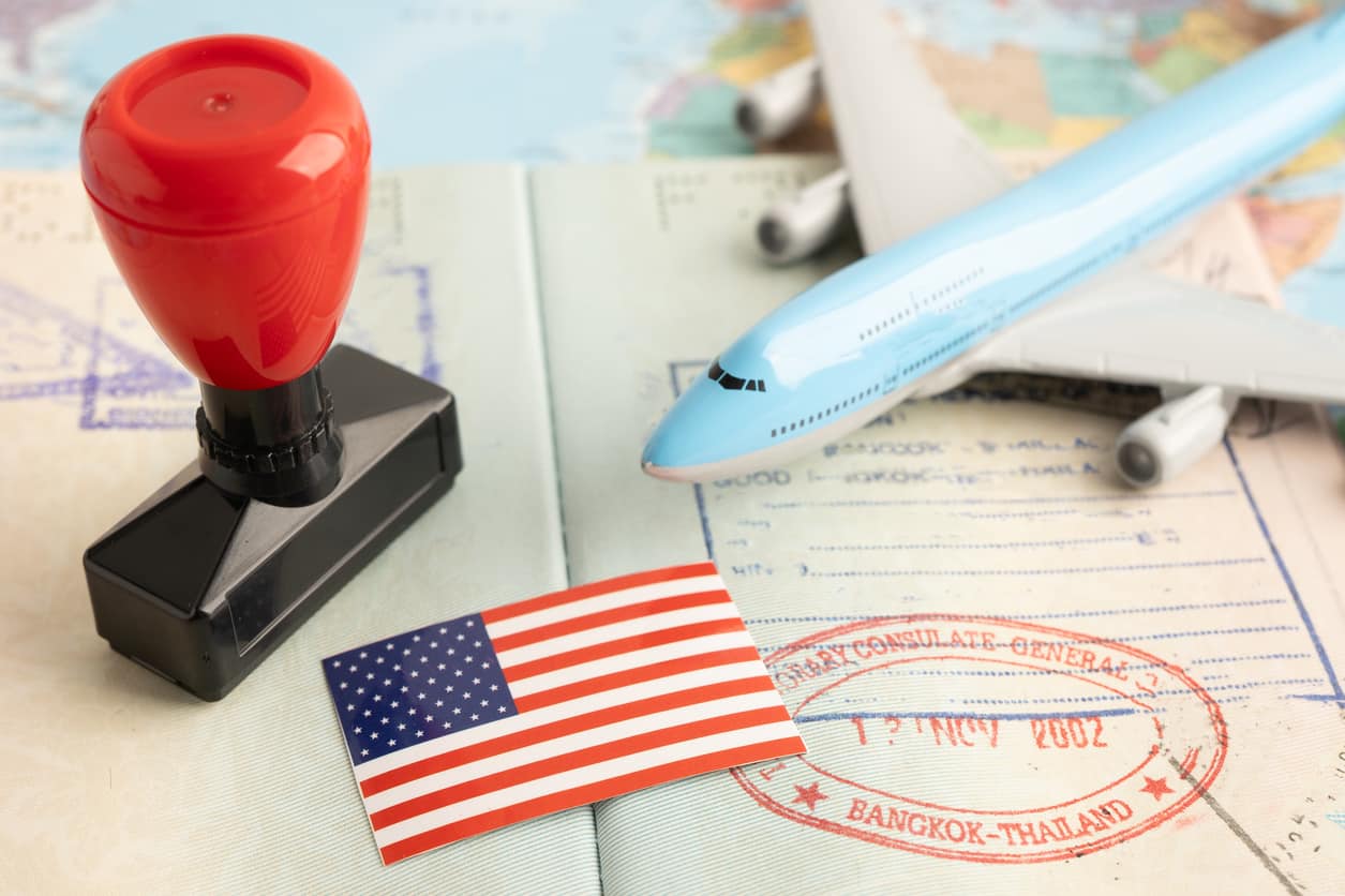 The image shows a U.S. visa with a red stamp, a toy airplane, and an American flag placed on top of a passport.
